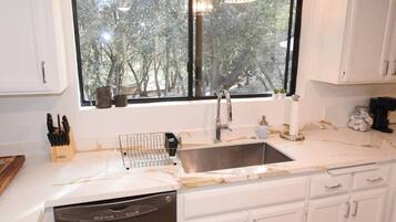 Una cocina con un lavamanos doble, armarios blancos, lavavajillas y una ventana con vistas a árboles.