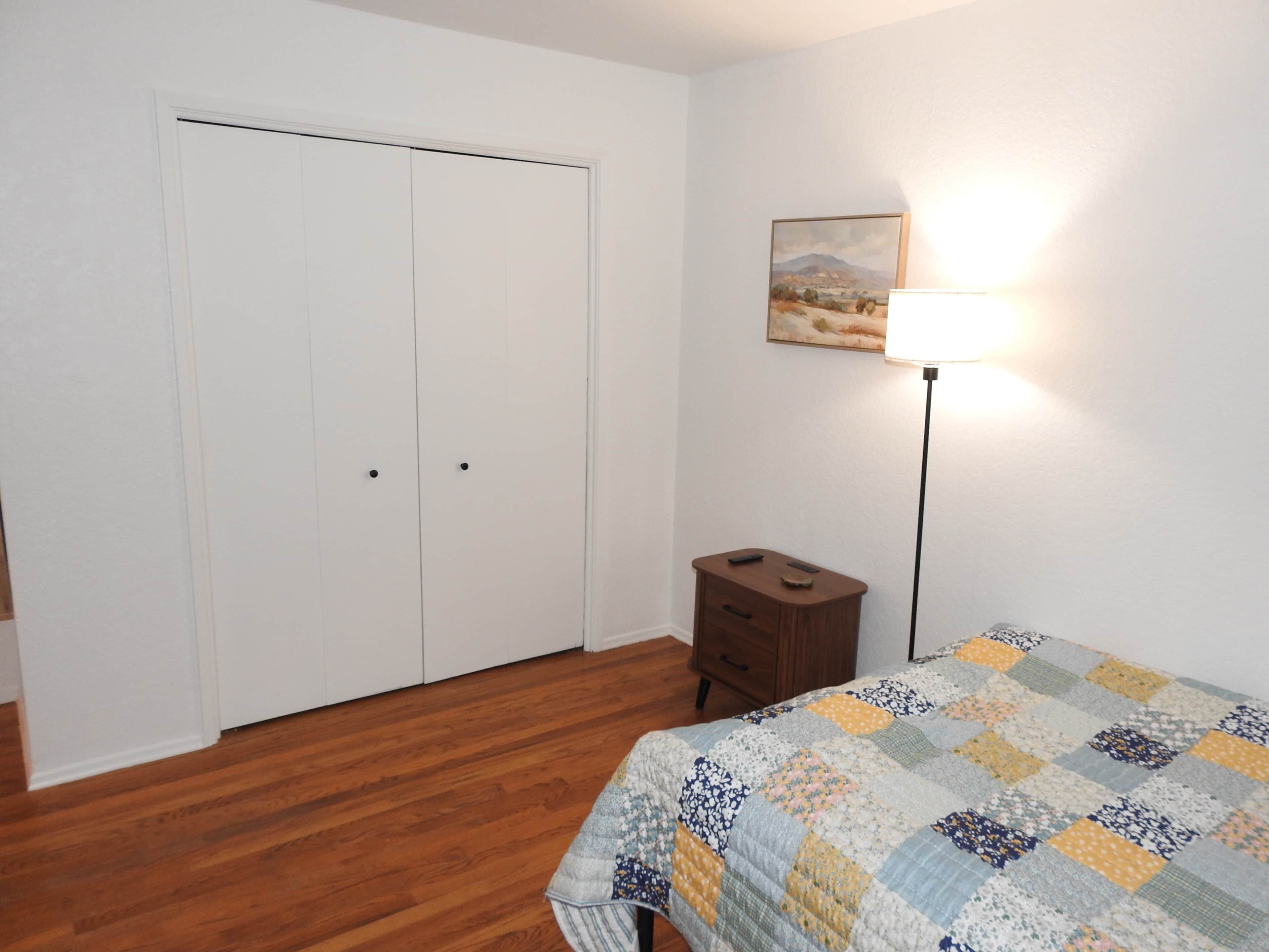 A bedroom with a wooden floor, a bed with a quilt, a nightstand, a lamp, and a door.
