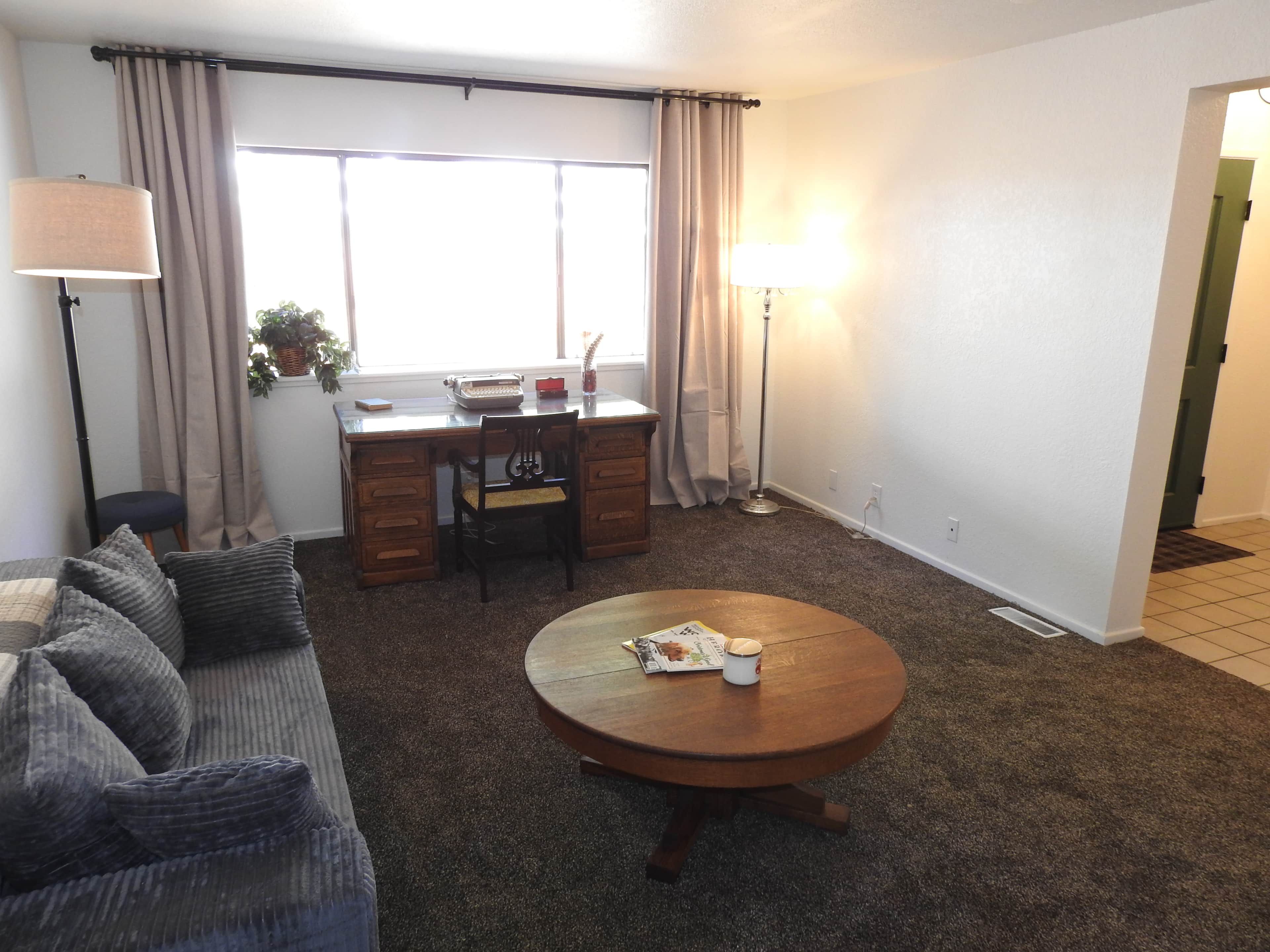 Sala de estar com sofá, mesa de centro redonda, escrivaninha com cadeira, luminária e janela com cortinas.