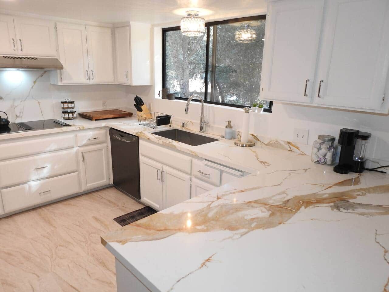 Uma cozinha moderna com armários brancos, bancada de mármore e janela com vista para árvores.