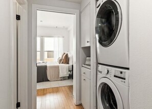 A laundry room with a front-loading washing machine and a dryer, a bed with a white headboard and pillows, and a window with curtains. - 2-bedroom apartment in Brentwood. The den/office is ideal for working from home (Los Angeles)