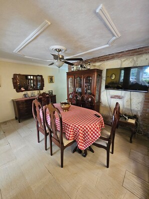 Un coin repas avec une nappe à carreaux, des chaises en bois, un ventilateur de plafond, un téléviseur fixé au mur et un mur en briques.