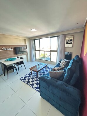 A living room with a blue sofa, a coffee table, a dining table with chairs, and a large window with curtains. - Melhores Flats - Divino, pertinho do mar (João Pessoa)