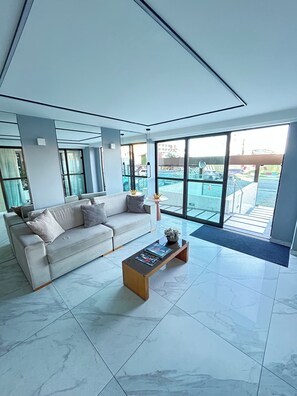 A modern living room with a sofa, coffee table, and large windows overlooking a swimming pool. - Melhores Flats - Divino, pertinho do mar (João Pessoa)