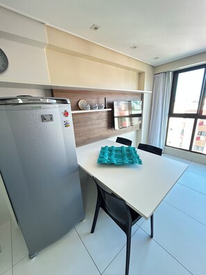 A modern kitchen with a refrigerator, a white table, and a window with curtains. - Melhores Flats - Divino, pertinho do mar (João Pessoa)
