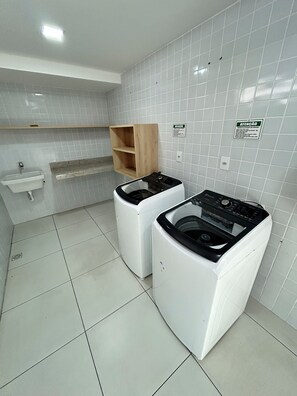 A laundry room with two front-loading washing machines, a sink, and a wooden shelf. - Melhores Flats - Divino, pertinho do mar (João Pessoa)