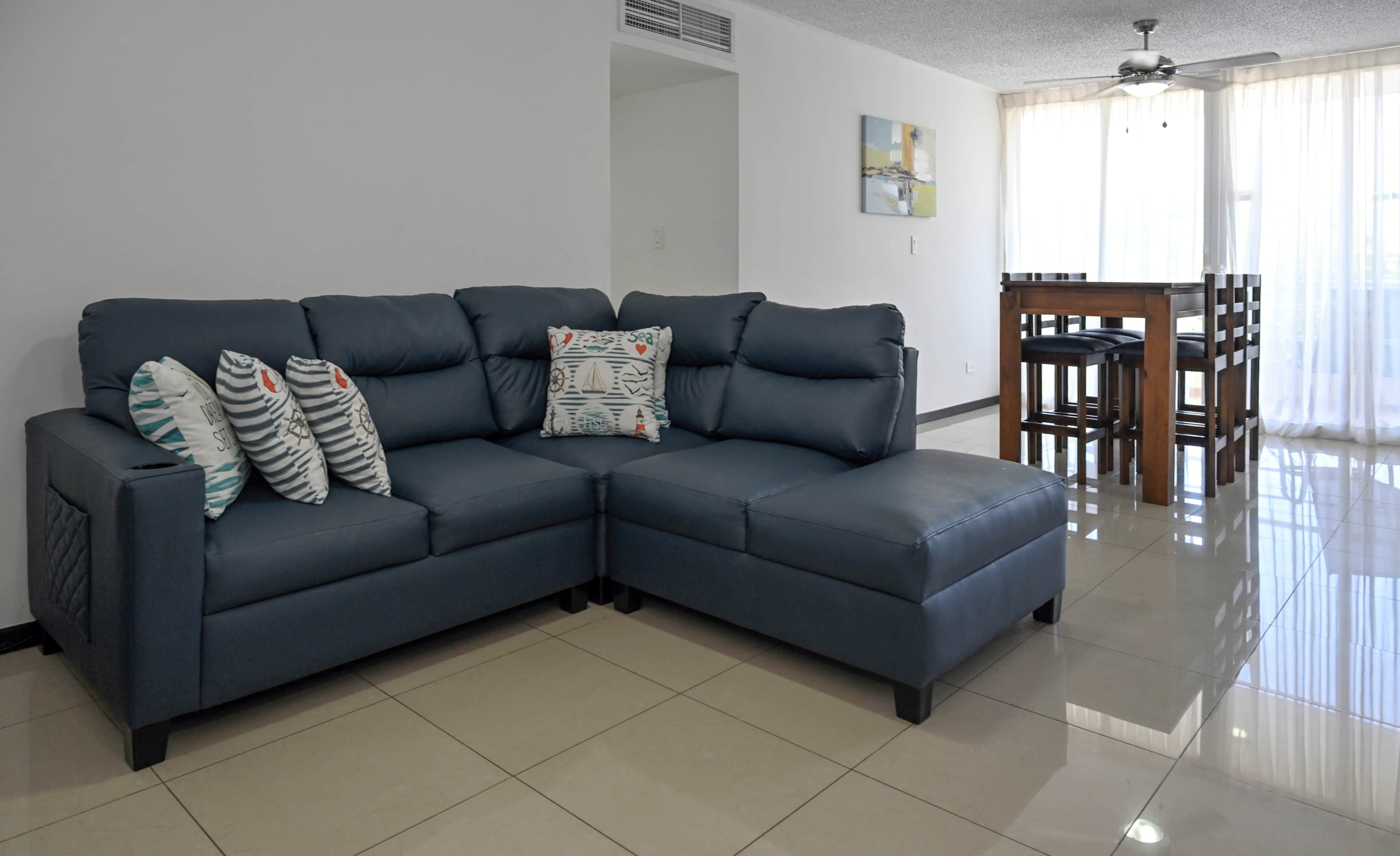 A modern living room with a dark blue sectional sofa, a dining table with chairs, and a ceiling fan.
