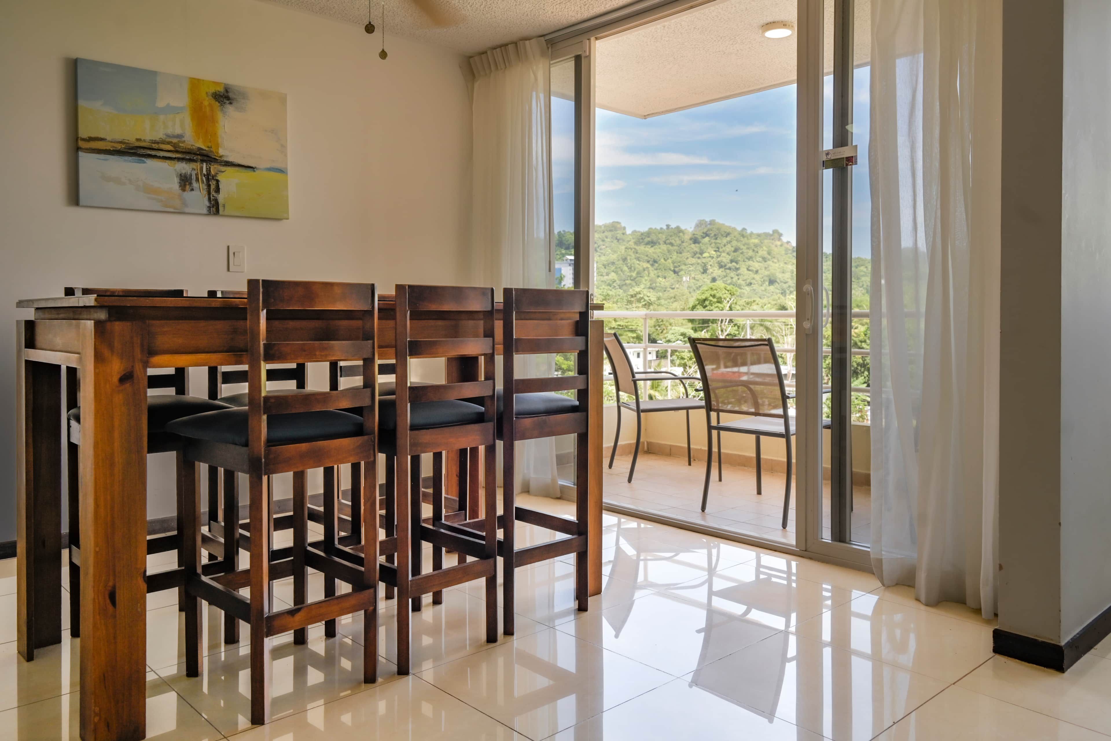 A dining area with a wooden table and chairs, a painting on the wall, and a view of a green landscape through the glass door.