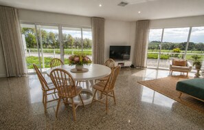 A spacious living room with a round dining table, comfortable seating, a flat-screen TV, and large windows offering a view of the outdoors.