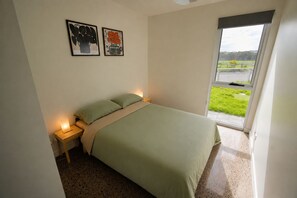 A bedroom with a bed, a nightstand, a lamp, and a window with a view of a grassy area.