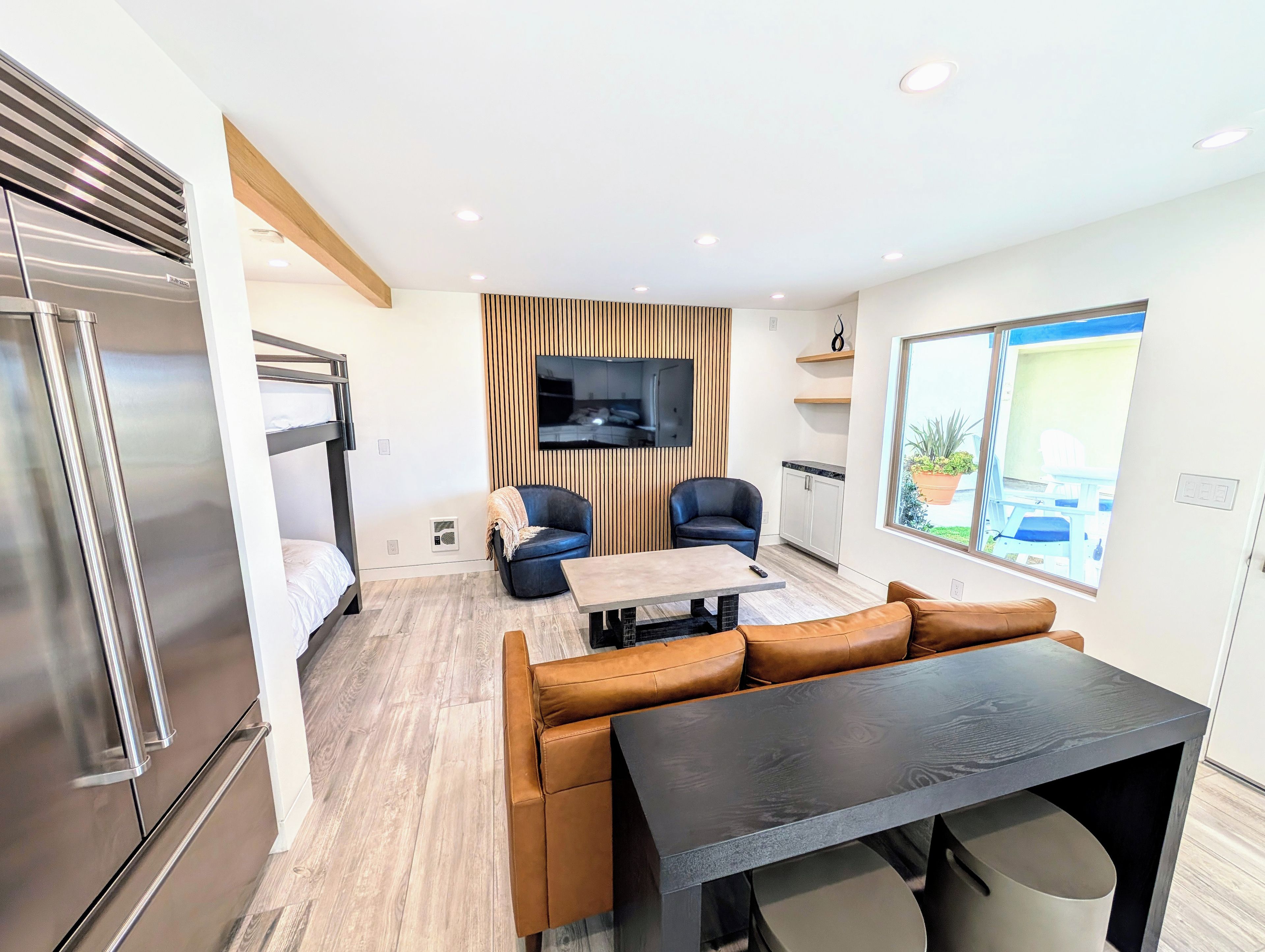 A modern interior with a bunk bed, two leather sofas, a dining table, and a TV mounted on a wooden panel wall.