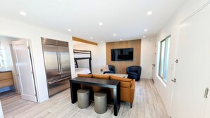 A compact living space with a kitchenette, a bunk bed, a seating area with a table, and a flat-screen TV mounted on the wall. - Beach Front - Studio Condo, Luxury Finishes, Walk to Everything, 440 sq ft, WIFI (Huntington Beach)