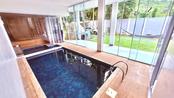 A modern indoor swimming pool with clear glass walls, a wooden deck, and a view of the outdoors.