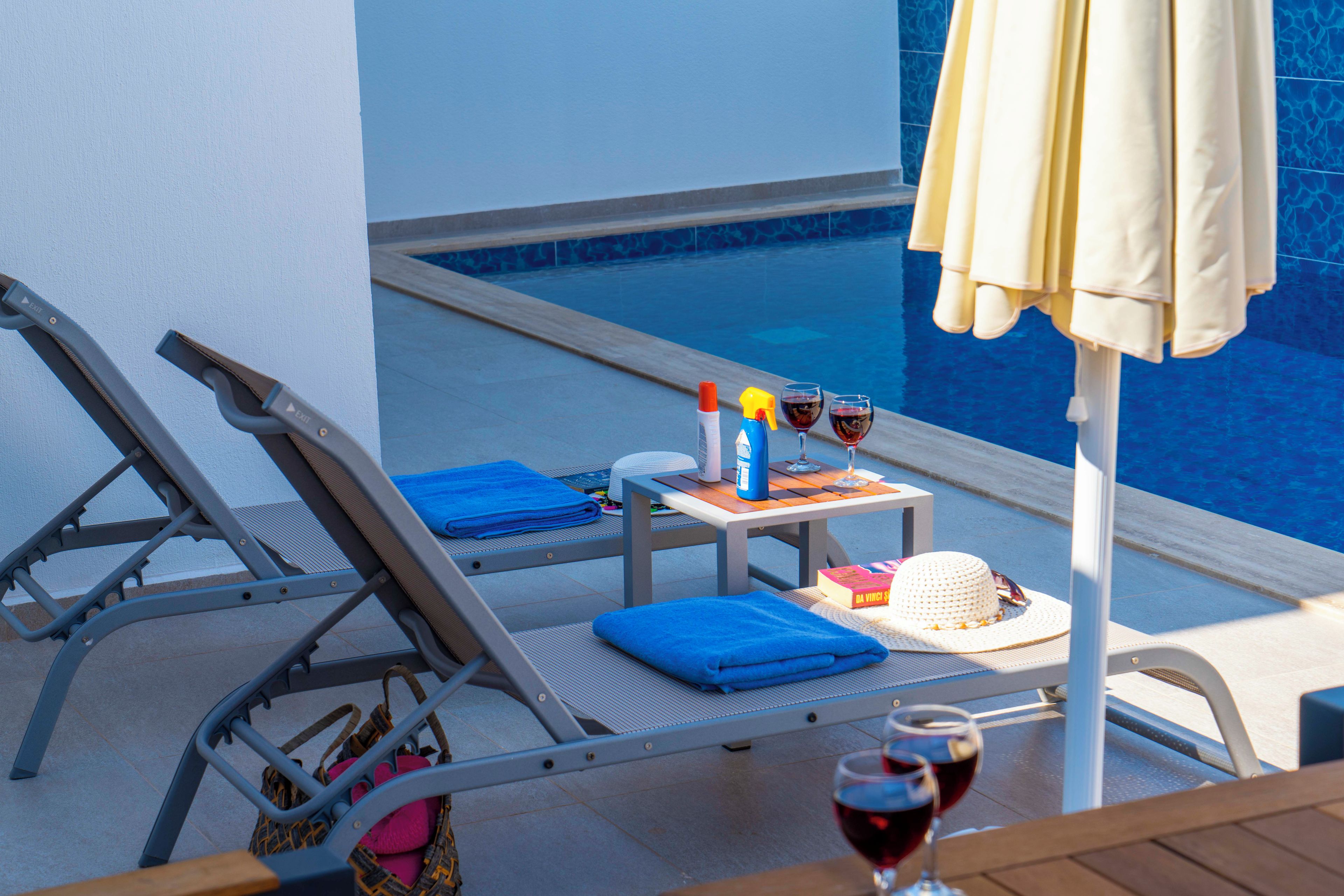 Área de descanso à beira da piscina com mesa, duas espreguiçadeiras, guarda-sol e uma garrafa de vinho.