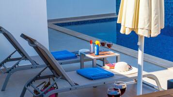 Área de descanso à beira da piscina com mesa, duas espreguiçadeiras, guarda-sol e uma garrafa de vinho.