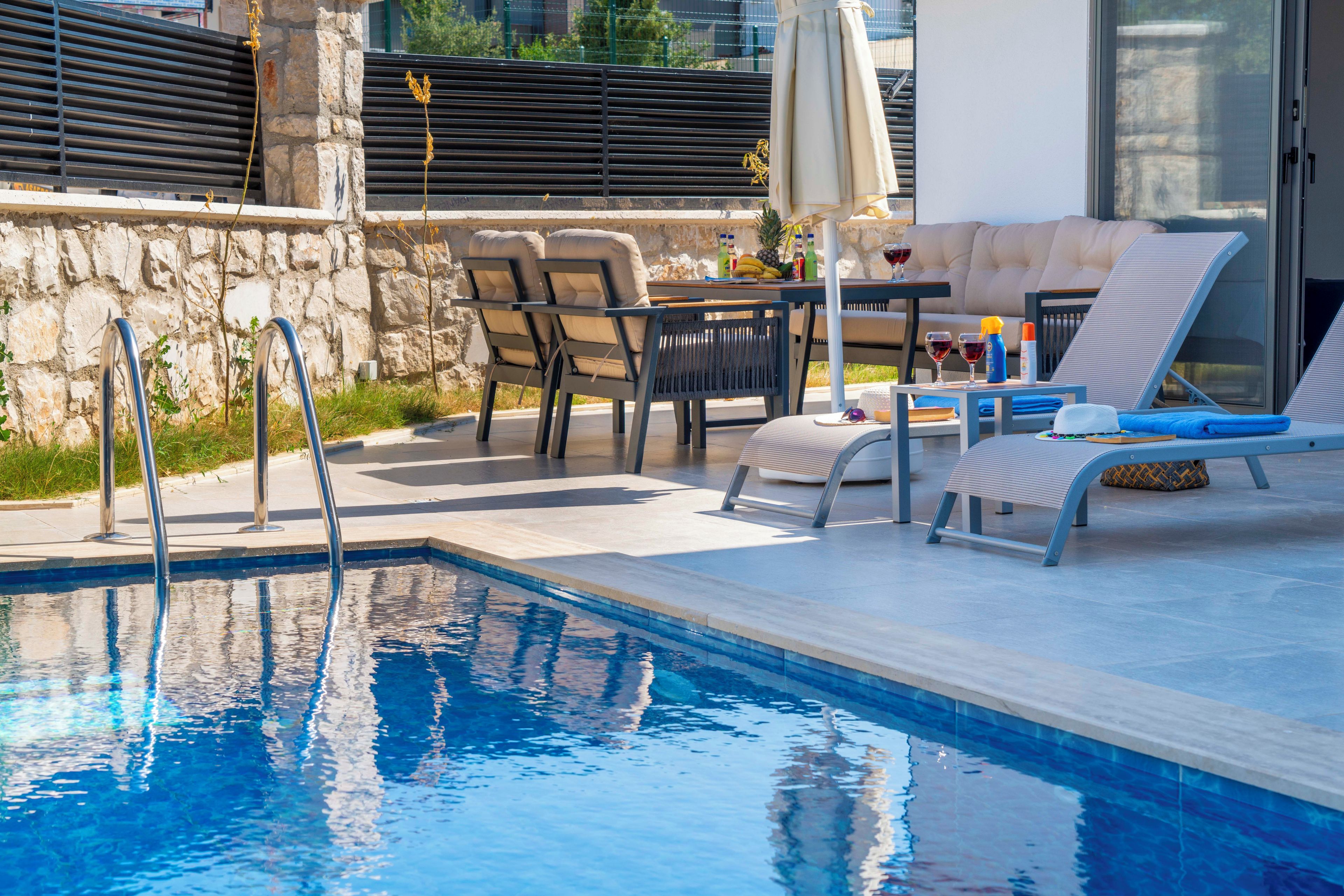 Una piscina con sillones de descanso y una mesa con bebidas, rodeada de muros de piedra y una zona de estar techada.