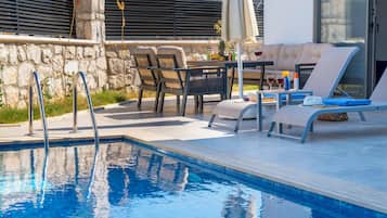 Una piscina con sillones de descanso y una mesa con bebidas, rodeada de muros de piedra y una zona de estar techada.