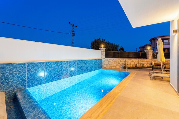 A swimming pool with clear blue tiles, a stone wall, and a wooden deck with lounge chairs.