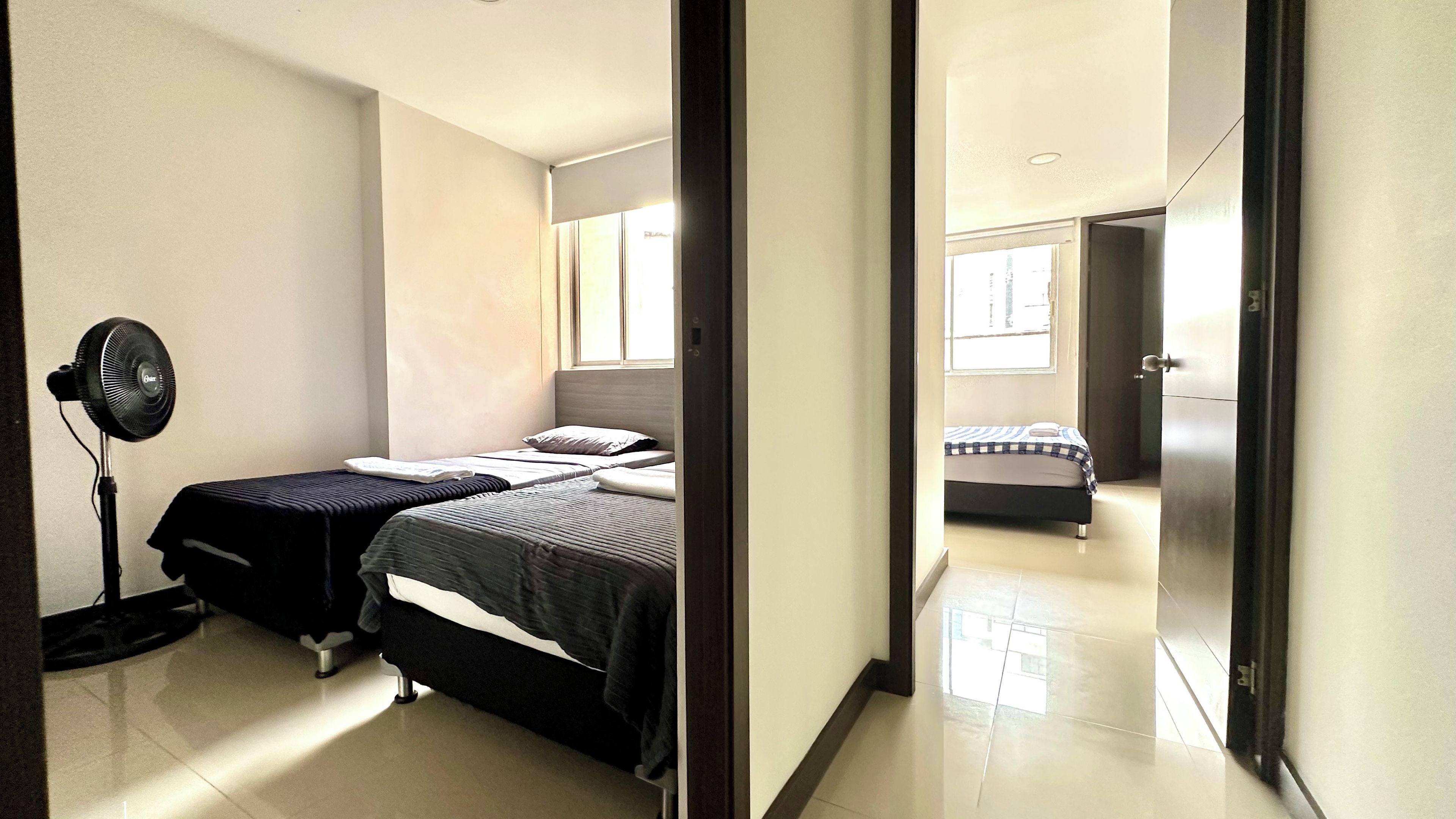Un dormitorio con dos camas, un ventilador de pie y vista a otra habitación a través de una puerta abierta.