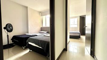 Un dormitorio con dos camas, un ventilador de pie y vista a otra habitación a través de una puerta abierta.