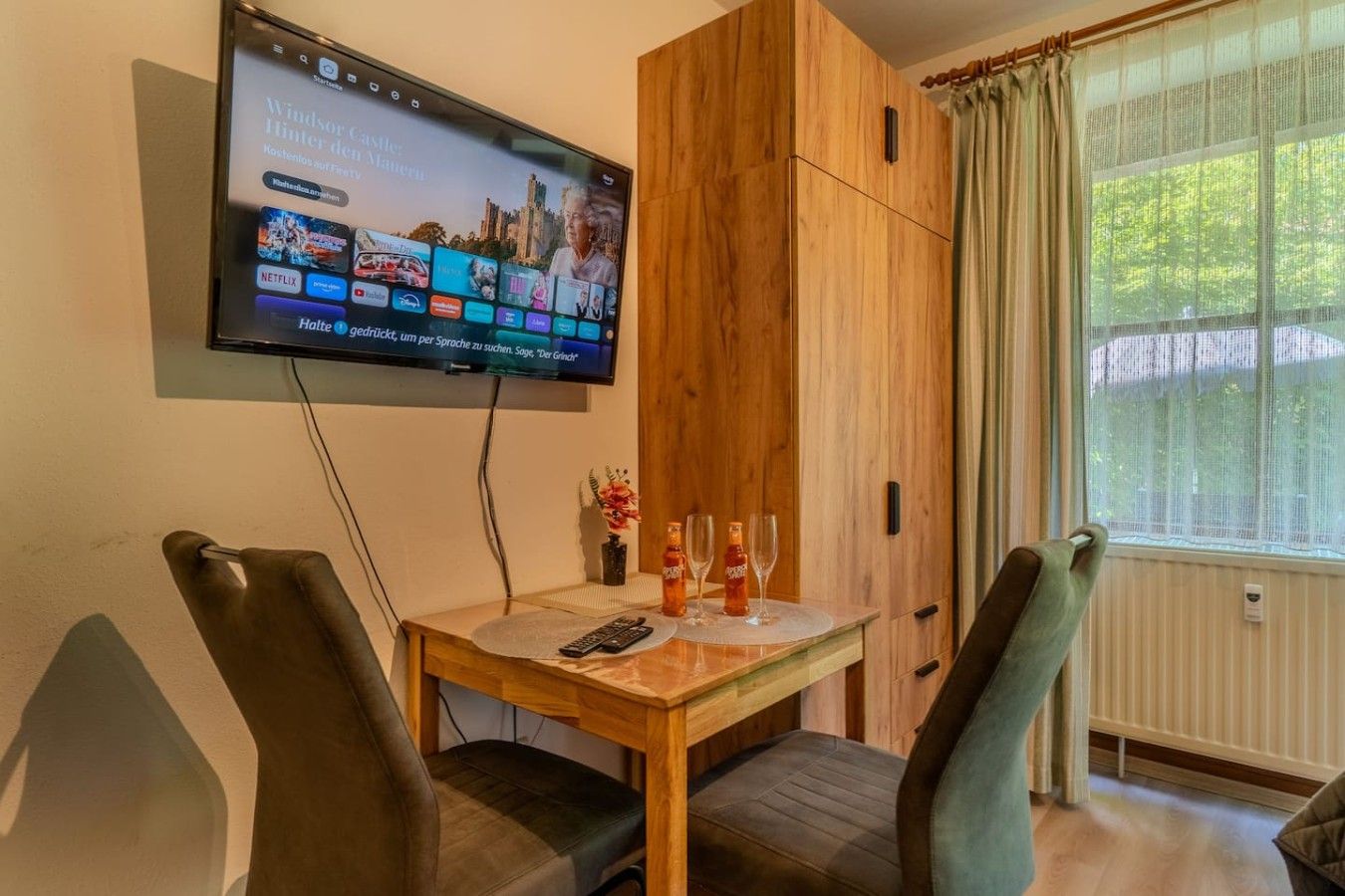 Ein Zimmer mit einem Holzkasten, einem an der Wand montierten Fernseher, einem Esstisch mit Stühlen und einem fenster mit Vorhängen.