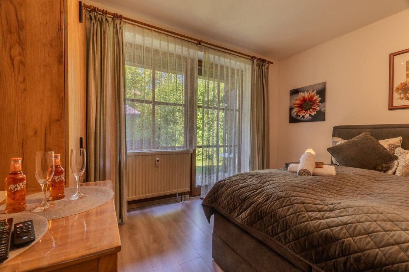 A bedroom with a large bed, a bedside table with a bottle and glasses, a radiator, and a window with curtains.