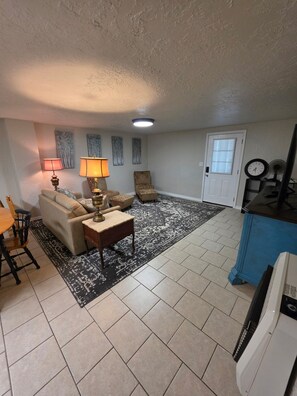A living room with a sofa, two chairs, a coffee table, a patterned rug, a door, and a television. - The Hidden Gem: Cozy Blue Cottage Sleeps 6, 10 min. to OU . Pet friendly (Noble)