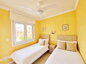 Ein Zimmer mit zwei Betten, einem Deckenventilator, einem Fenster und einem Nachttisch.