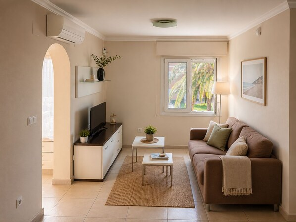 A living room with a sofa, TV, small table, and a window with a view of palm trees. - Oliva Nova MET & Beach - Apartment for 4 people, 400 m from the beach and 50 MET from the Costa Blanca (VC)