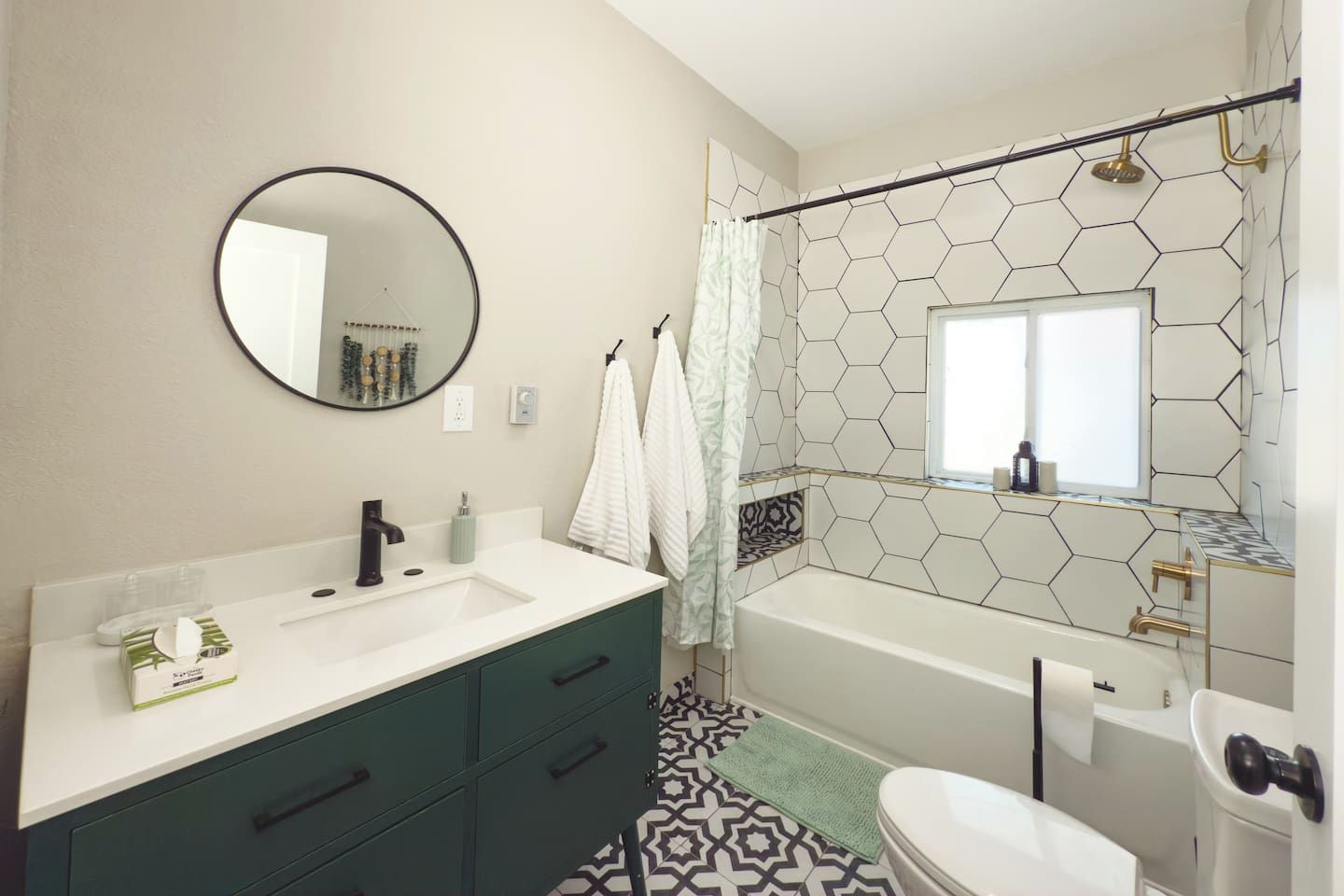 A bathroom with a white sink, dark green cabinets, a round mirror, a shower with a hexagonal tile pattern, and a toilet.