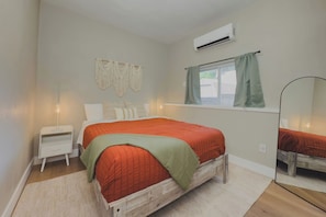 A bedroom with a wooden bed, a nightstand, a bedside lamp, a macrame wall hanging, an air conditioning unit, a window with curtains, and a mirror. - Travelers Dream - 2 Homes 1 Stay (Colorado Springs)