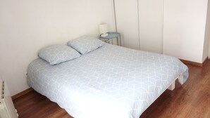 Quarto com cama, duas almofadas, mesa de cabeceira com abajur e uma cadeira azul pequena.