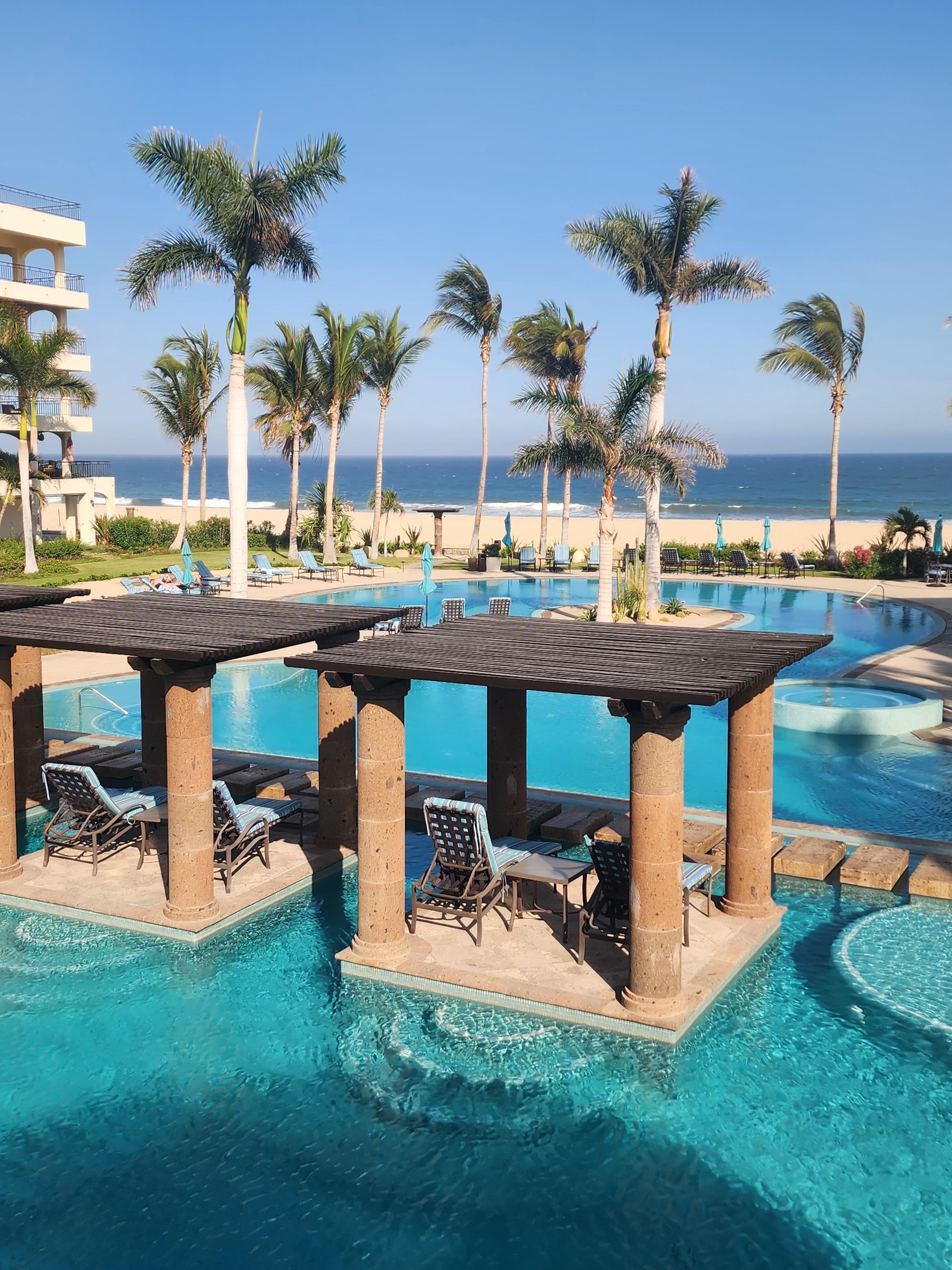 Zona de piscina en un resort con sillones de playa y sombrillas, vista despejada del océano y un edificio con balcones.