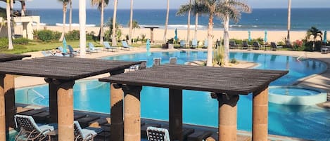 Zona de piscina en un resort con sillones de playa y sombrillas, vista despejada del océano y un edificio con balcones.