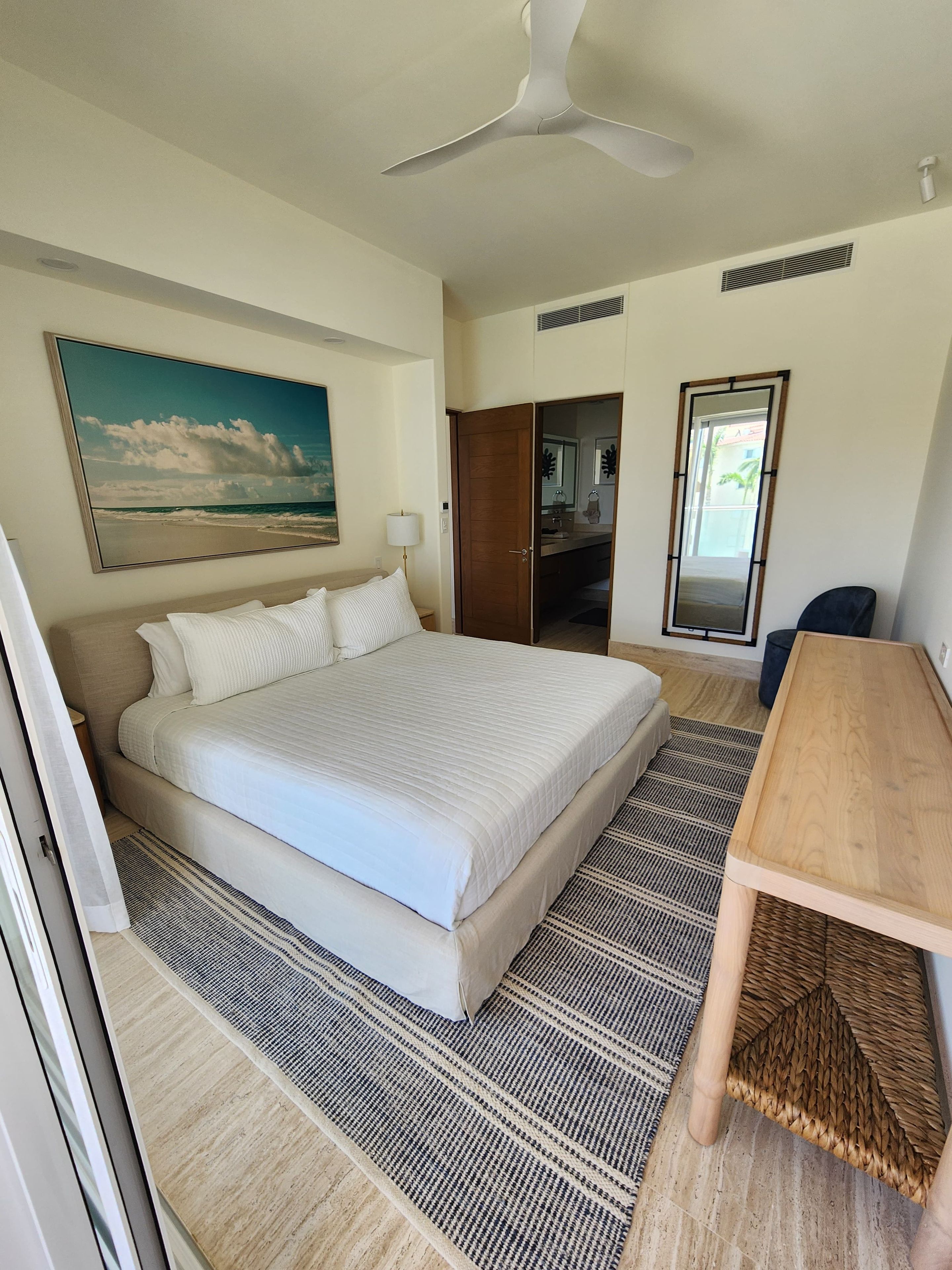 Habitación de hotel con cama, escritorio, ventilador de techo, una pintura de playa y vistas al exterior.