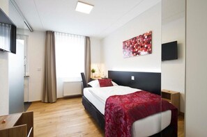 Habitación de hotel con cama, escritorio, silla, televisor y ventana con cortinas.