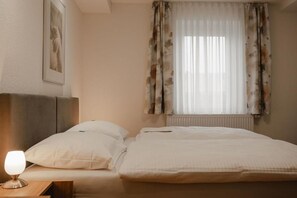 Quarto com cama, almofadas, mesa de cabeceira com abajur, quadro na parede e janela com cortinas.