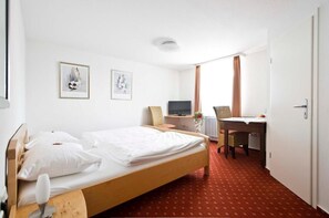 Habitación de hotel con una cama individual, un escritorio, una silla, un televisor y dos cuadros en la pared.