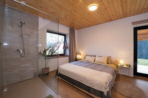 A modern bedroom with a glass shower enclosure, a large bed with multiple pillows, and a view of the outdoors through a window.