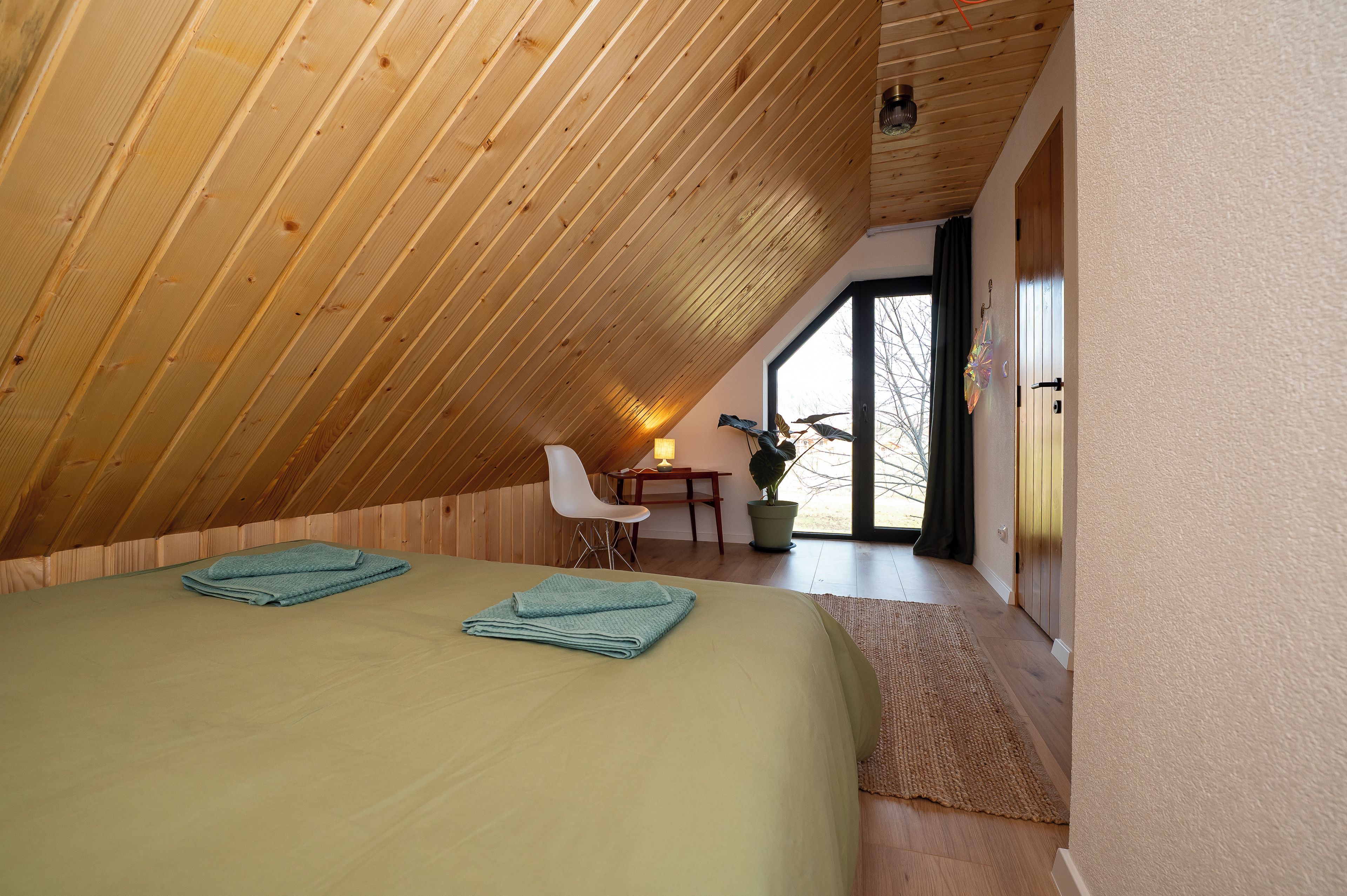 Een slaapkamer met een schuine kap, een bed met opgevouwen handdoeken, een witte stoel, een tafel met een lamp en een potplant.