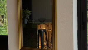 Ein großer Spiegel mit goldener Rahmung an einer strukturierten Wand, der einen Raum mit einem hölzernen Hocker und einem Blumentopf widerspiegelt.