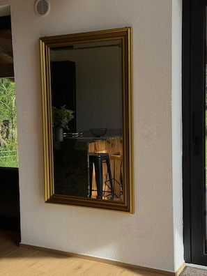 Un espejo grande con marco dorado sobre una pared con textura, que refleja una habitación con un taburete de madera y una planta en maceta.