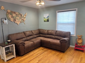 Un salon avec un canapé d’angle, un plancher en bois, une carte des États-Unis accrochée au mur, une fenêtre avec des stores et une petite table en bois.