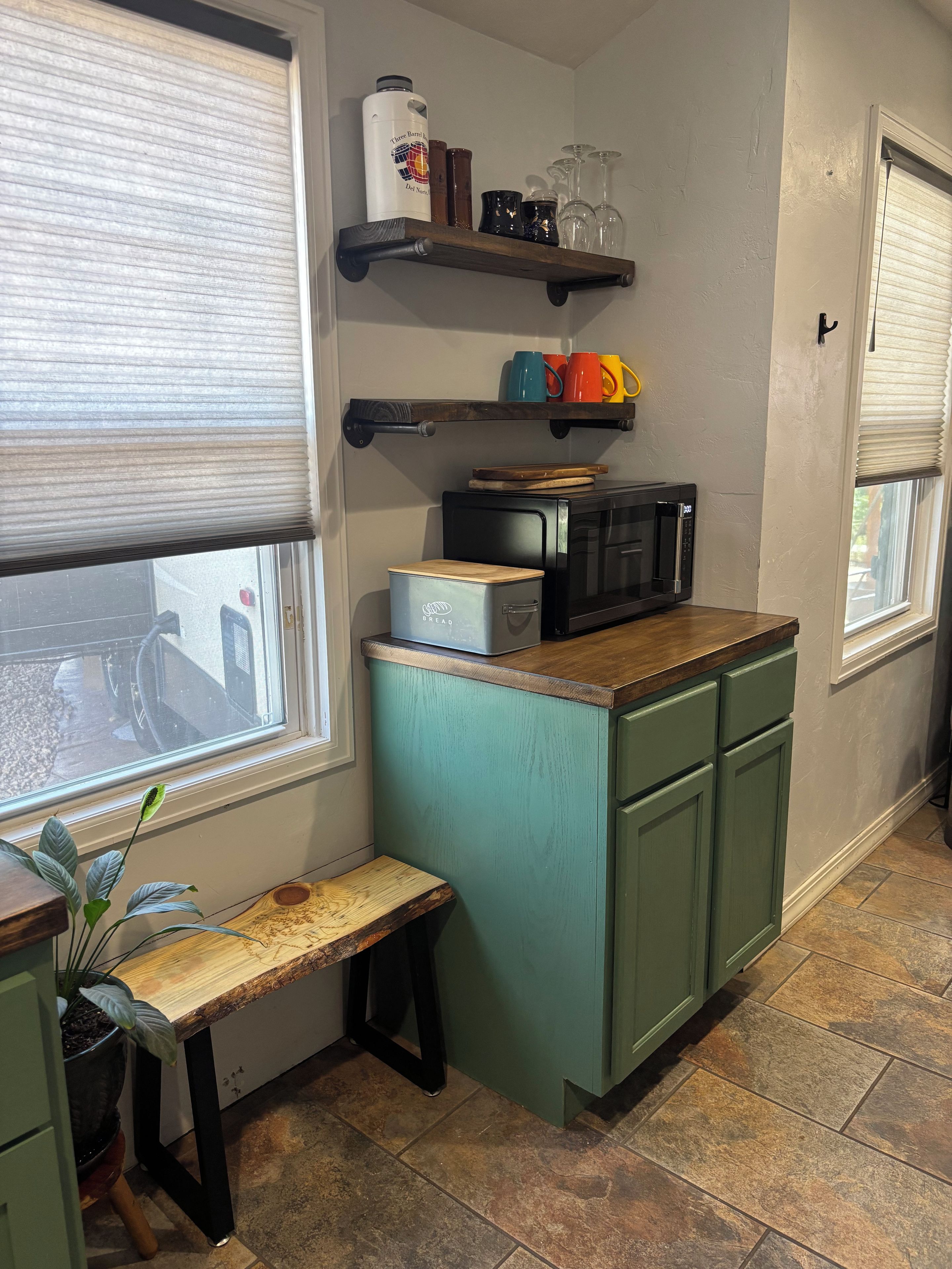 A kitchen with a microwave, a bread box, and a shelf with mugs and glasses.