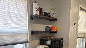 Una cocina con microondas, panera y estante con tazas y vasos.