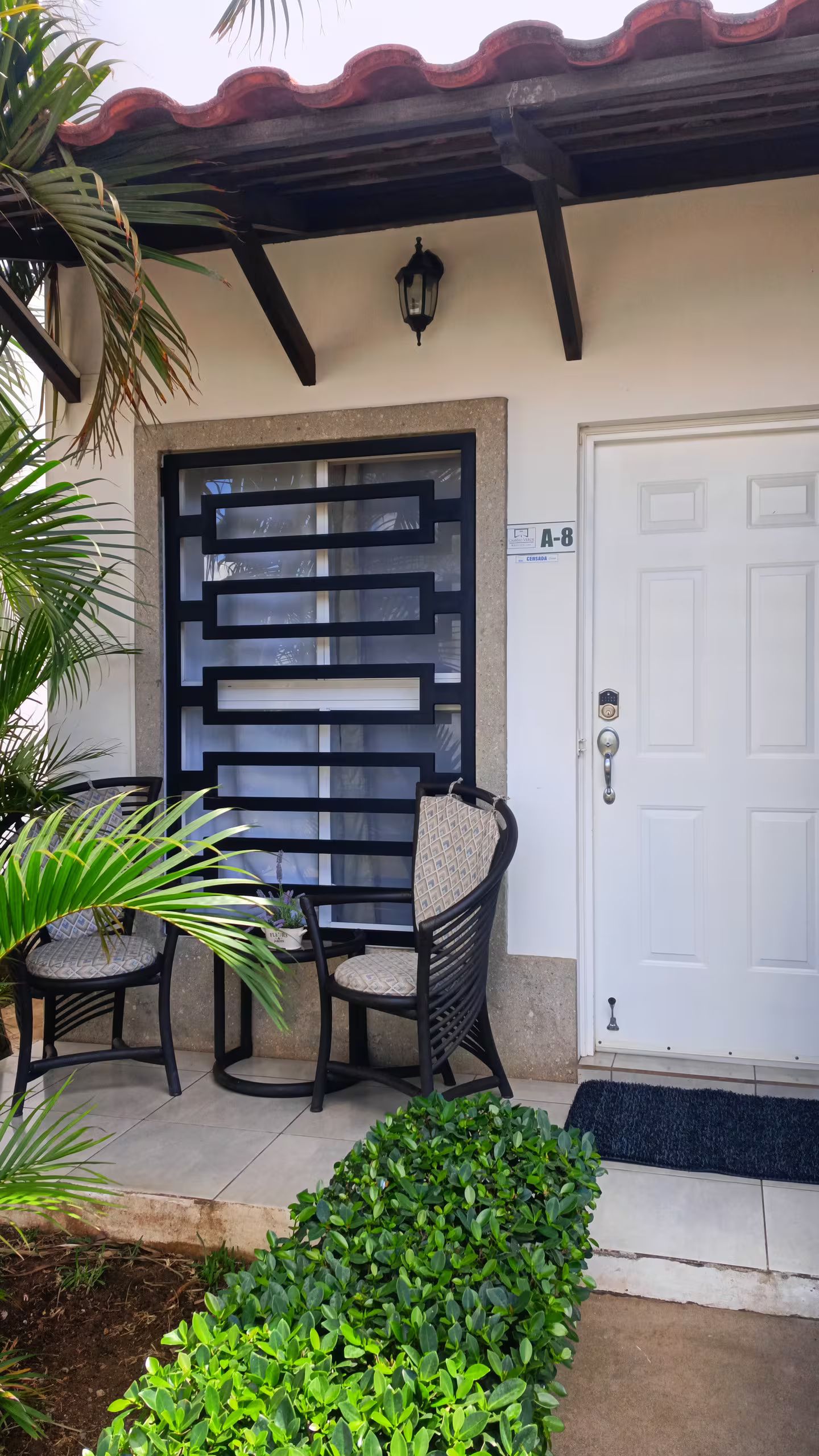 Entrada de un edificio con una puerta blanca, un portón de metal negro, dos sillas de mimbre y una pequeña planta en maceta.