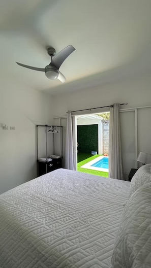 A bedroom with a bed, a ceiling fan, a nightstand, a hanging light, and a view of a swimming pool through an open door.