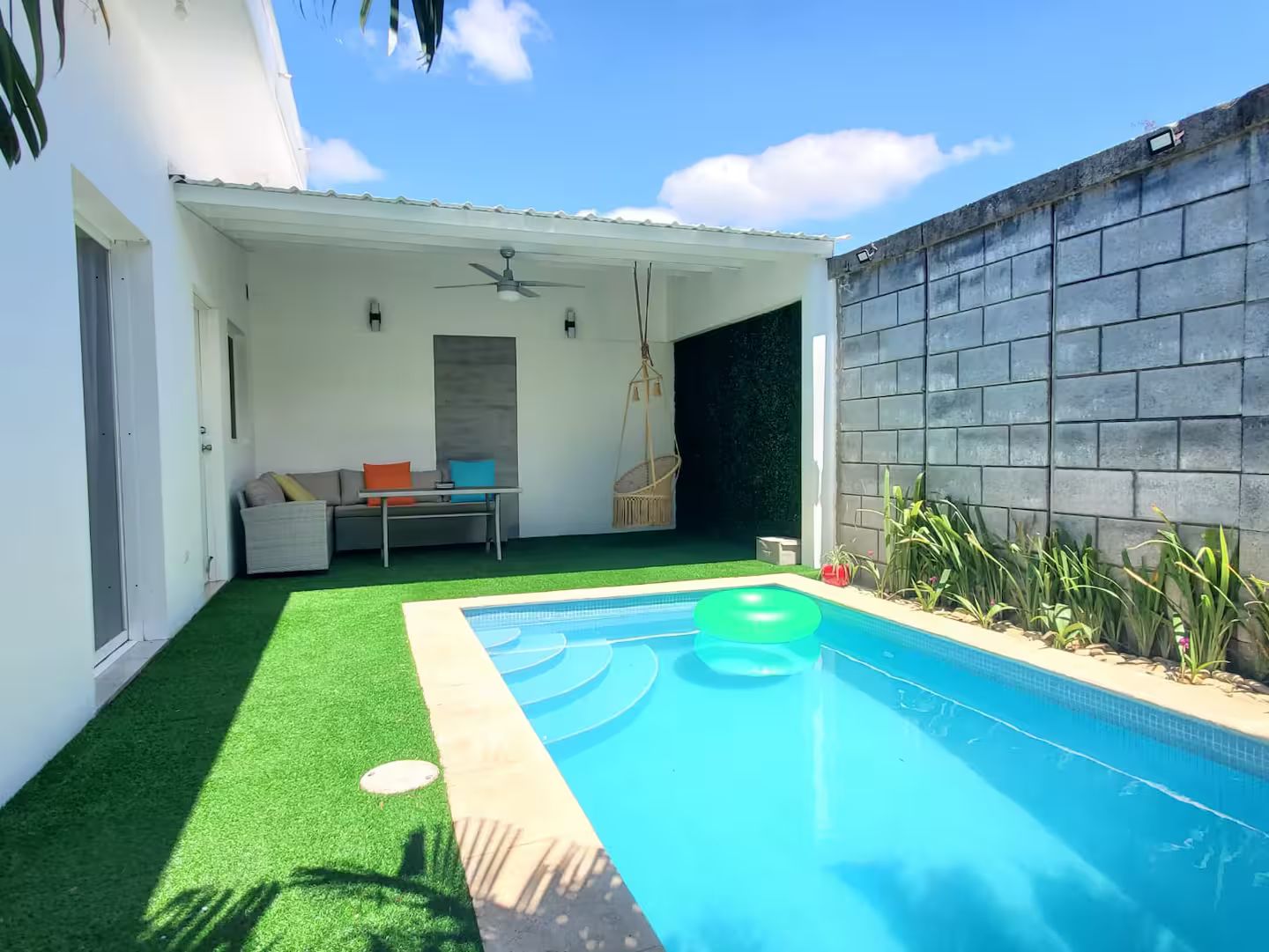 Una piscina con césped artificial, un área de estar con sofá y mesa, y una silla colgante.