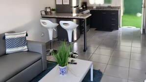 A modern living room with a sofa, a coffee table, and a kitchen area with a refrigerator, stove, and bar stools.