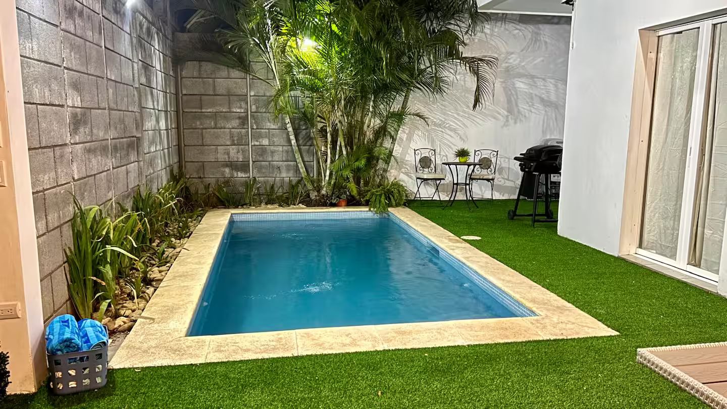 Piscina retangular com borda azulejada, rodeada por grama sintética, com uma parede de concreto de um lado e uma parede branca do outro.